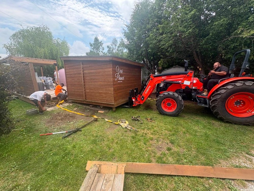 Orange tractor with a front loader lifting a large brown wood clad shed while two workers guide it, straps and pry bars on the grass